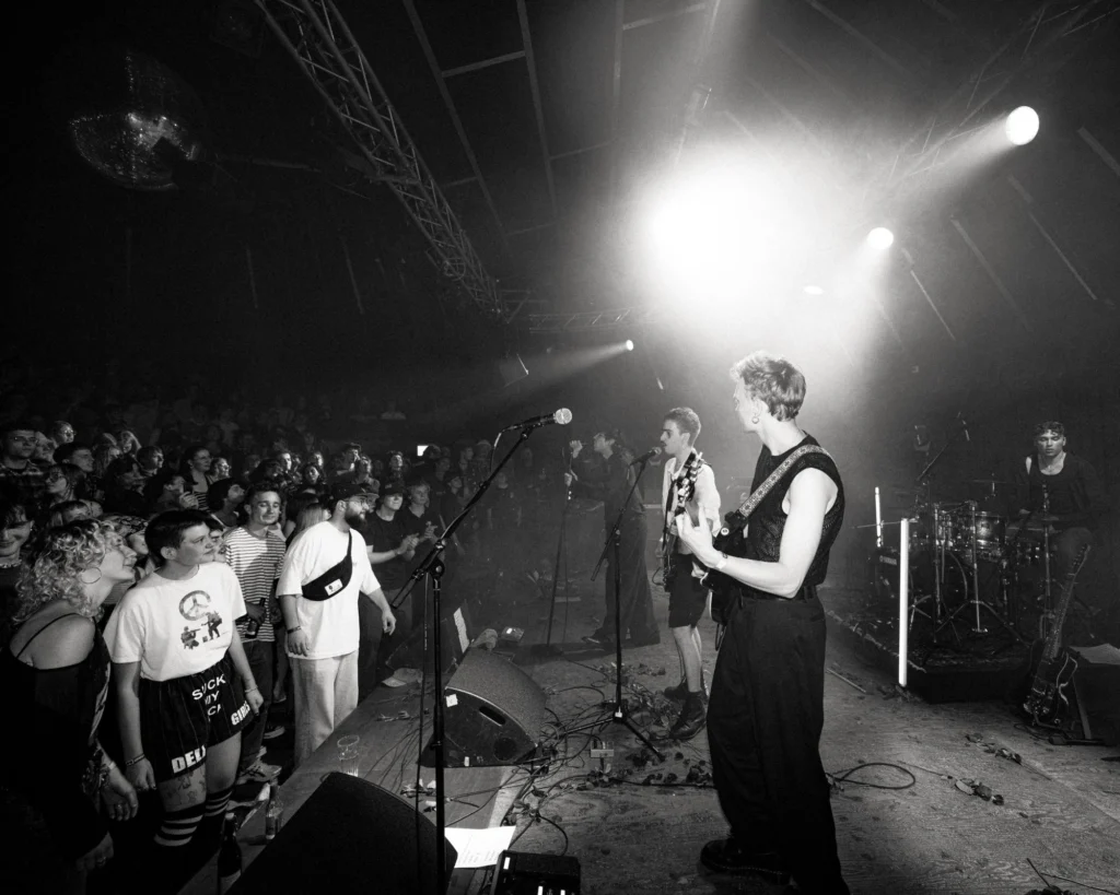 Schwarz-Weiss-Foto: Thierry Bill an der Gitarre auf einer grossen Bühne mit Band, im Scheinwerferlicht vor vollem Publikum