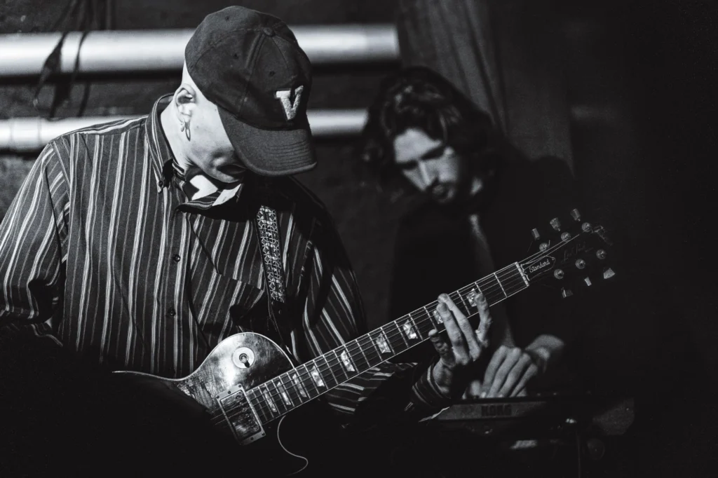 Thierry Bill mit Cap und gestreiftem Hemd spielt eine Les-Paul-Gitarre, im Hintergrund ein Keyboarder