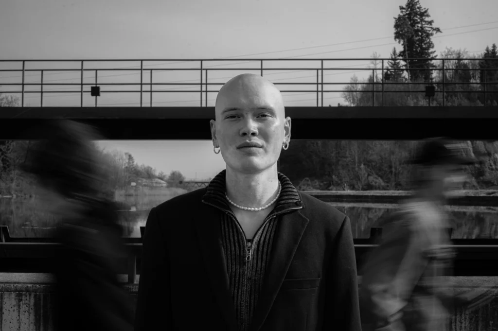 Schwarz-Weiss-Porträt von Thierry Bill mit Perlenkette vor einer Brücke, links und rechts verwischte Passanten